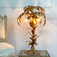 A decorative Hans Kogl table lamp shaped like a palm tree, made of metallic gold-toned metal leaves and trunk, with three glowing globe bulbs. It is placed on a glass-top side table against a pale wall, with part of a chair visible on the left.