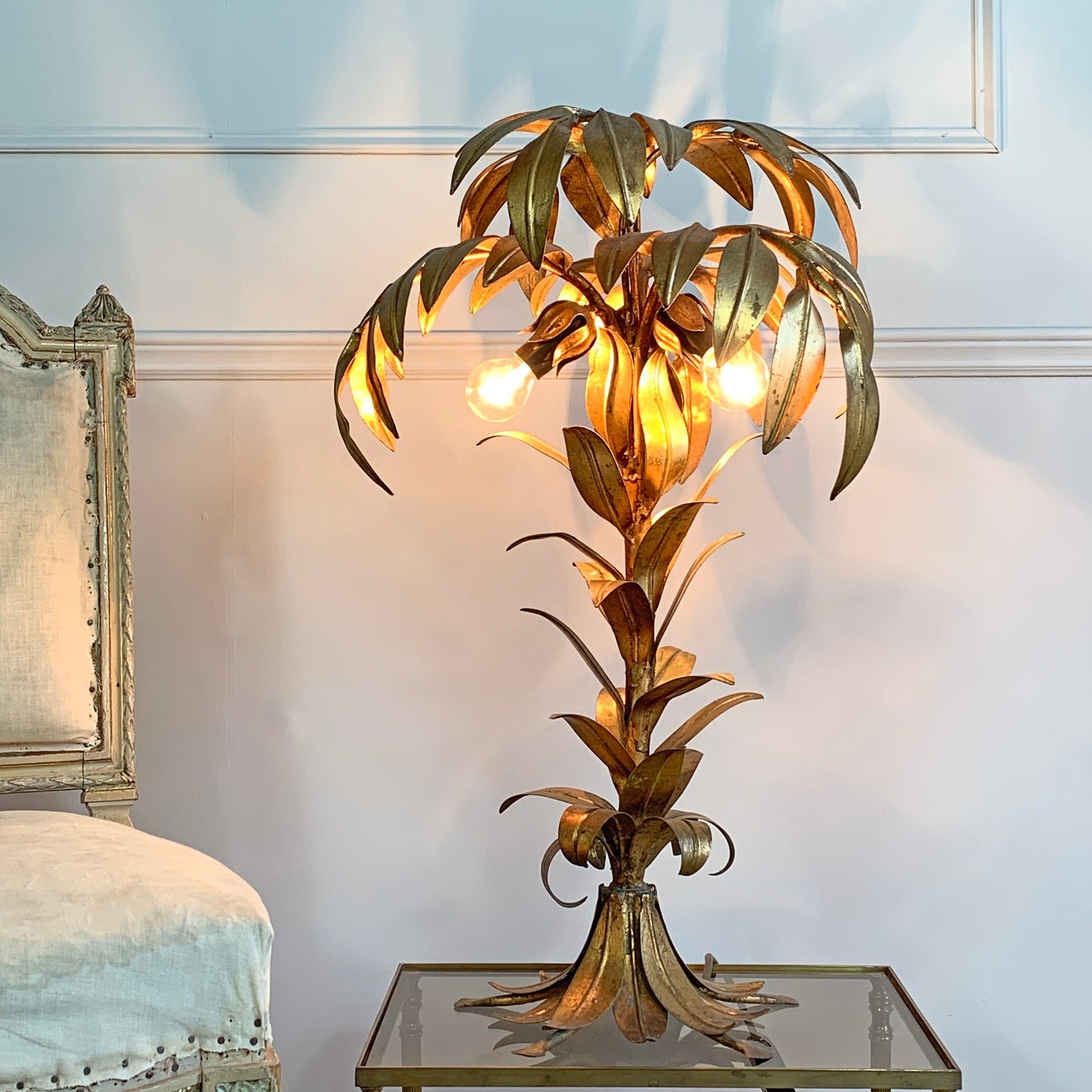 A decorative Hans Kogl table lamp shaped like a palm tree, made of metallic gold-toned metal leaves and trunk, with three glowing globe bulbs. It is placed on a glass-top side table against a pale wall, with part of a chair visible on the left.