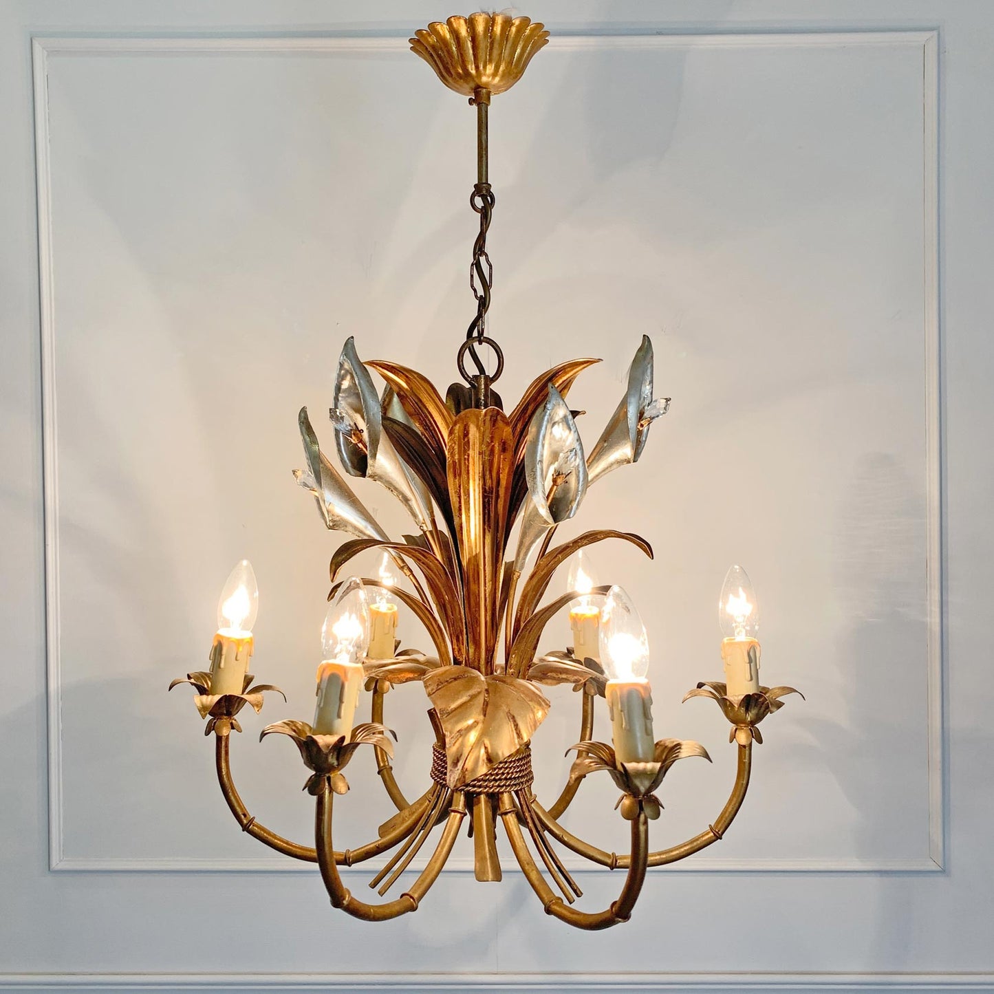 A decorative vintage hans kogl six-light chandelier with a warm gold and silver metallic finish, featuring sculpted calla lily and leaf elements and candle-style bulbs, shown hanging against a white paneled wall.