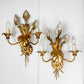 A pair of vintage hans kogl ornate gold wall sconces with leaf and floral motifs, each holding three candle-style bulbs with clear glass shades, mounted on a white wall.