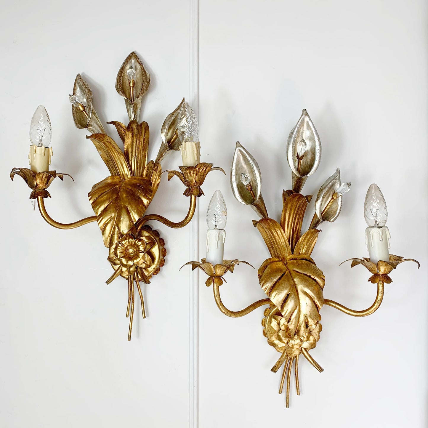 A pair of vintage hans kogl ornate gold wall sconces with leaf and floral motifs, each holding three candle-style bulbs with clear glass shades, mounted on a white wall.