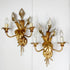 A pair of vintage hans kogl ornate gold wall sconces with leaf and floral motifs, each holding three candle-style bulbs with clear glass shades, mounted on a white wall.