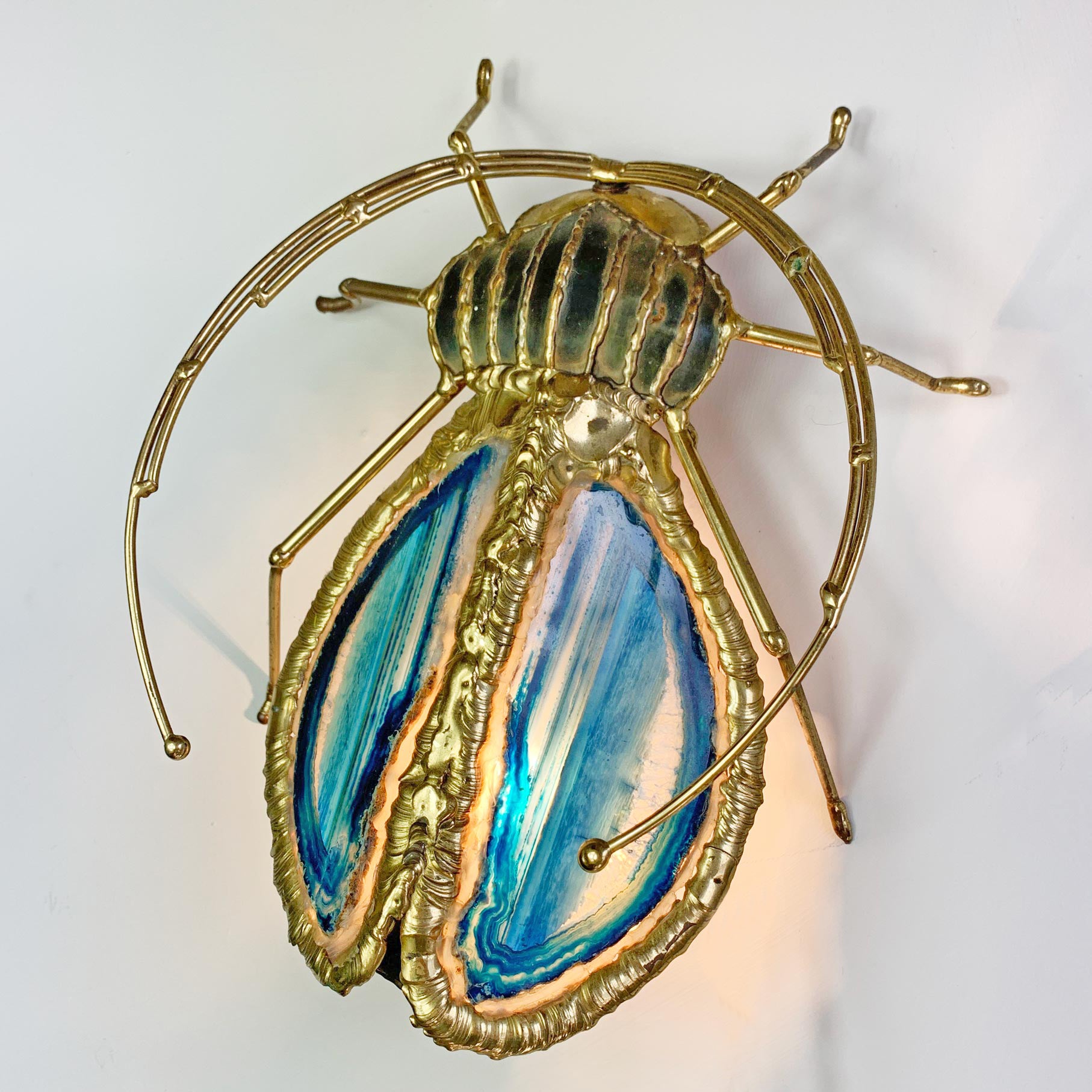 A Henri Fernandez Illuminated Gold Blue Agate Sculpture of a Longhorn Beetle beetle wall light with a gold metal frame and wings featuring blue, teal, and cream marbled patterns, shown against a plain white background.