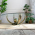 An oval glass-top coffee table with a sculptural base resembling an octopus in gold, bronze, and green tones, with a decorative striped sphere in the center, set on a beige rug in a styled interior.