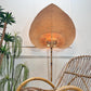 A large teardrop-shaped woven rattan wall light with a warm glowing light inside, styled in a room next to a wicker chair and potted plants against a white wall.