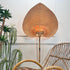 A large teardrop-shaped woven rattan wall light with a warm glowing light inside, styled in a room next to a wicker chair and potted plants against a white wall.