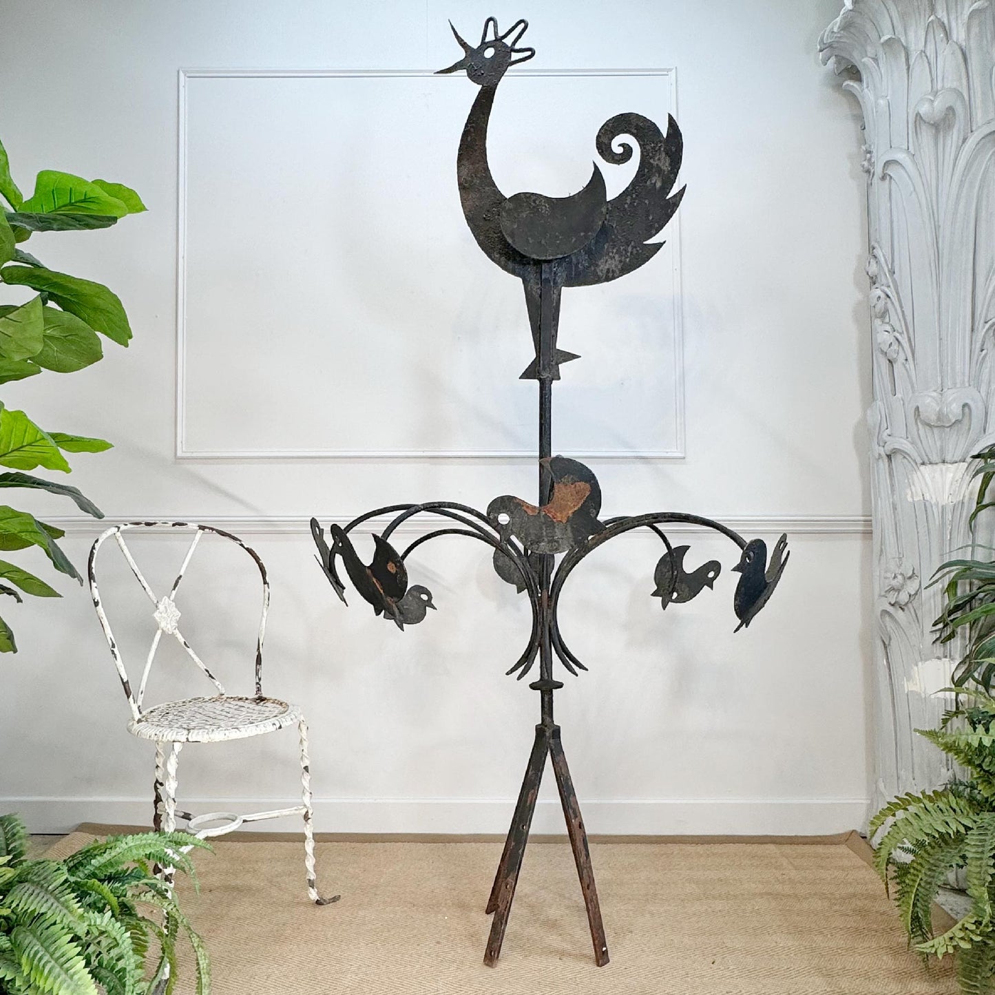 A rustic black metal garden statue shaped like a rooster with birds on curved branches, displayed in a styled room setting with a white chair, potted plants, and a white wall.