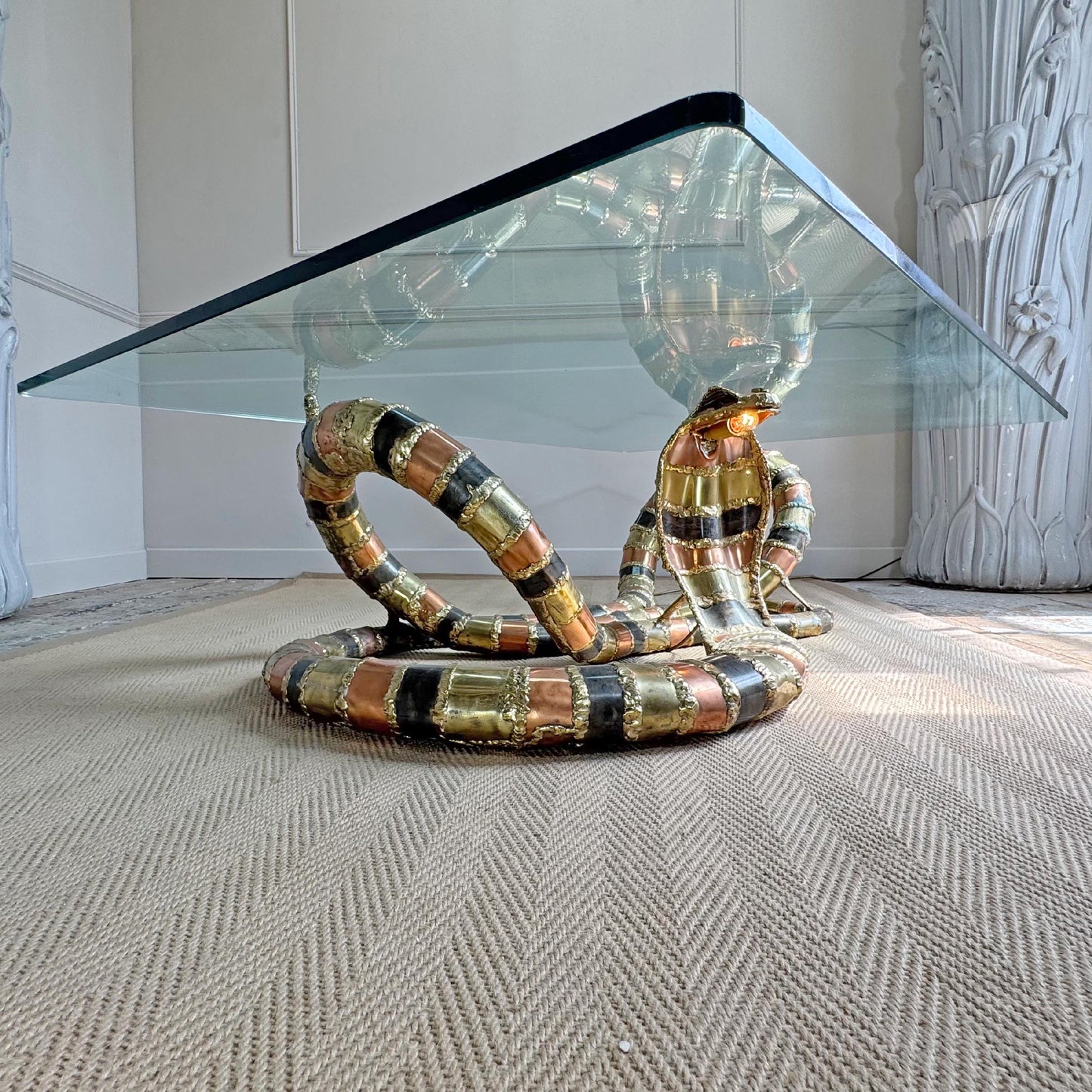 A modern Isabelle Faure snake-head coffee table with a clear glass rectangular top and a sculptural base coiled like a snake, featuring metallic gold, bronze, and black bands, set on a textured beige rug.