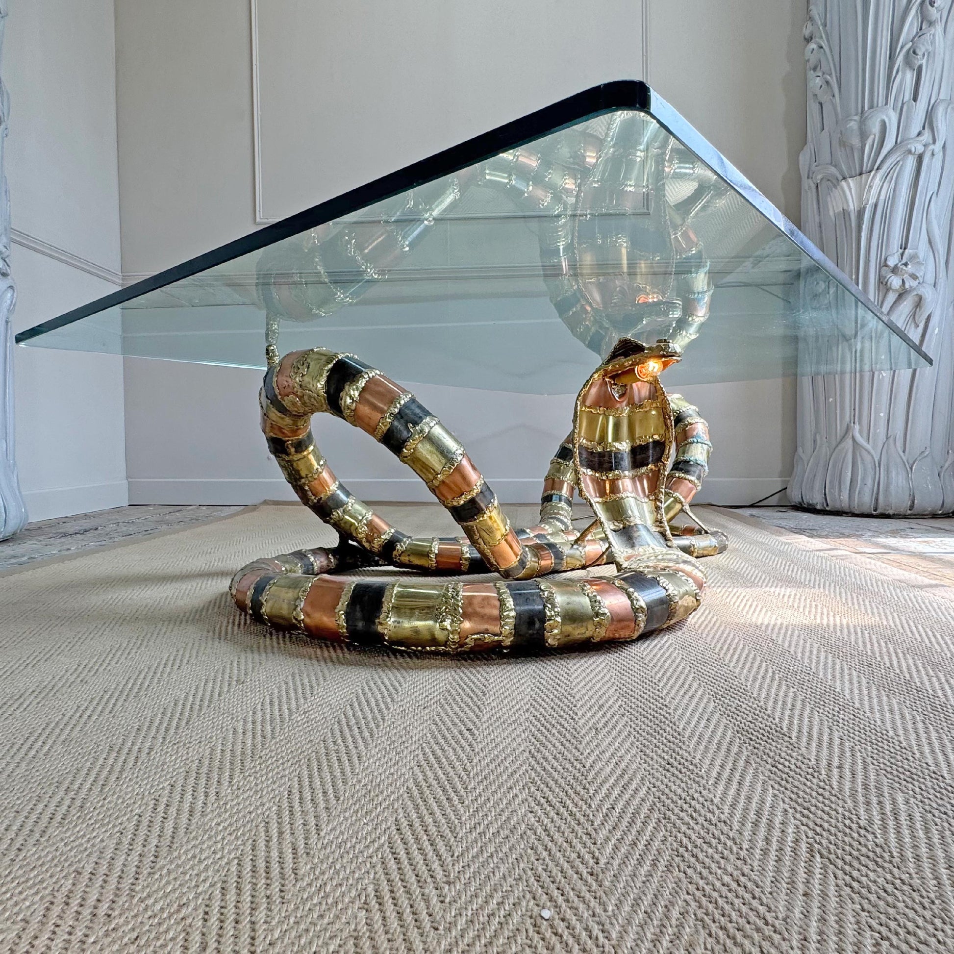 A modern Isabelle Faure snake-head coffee table with a clear glass rectangular top and a sculptural base coiled like a snake, featuring metallic gold, bronze, and black bands, set on a textured beige rug.