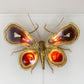 A decorative butterfly wall sculpture by Isabelle Faure,  made from gold-toned metal and polished agate slices, mounted on a white wall. The wings show layered natural agate patterns in brown, cream, and orange, with glowing warm light inside the lower wing sections.