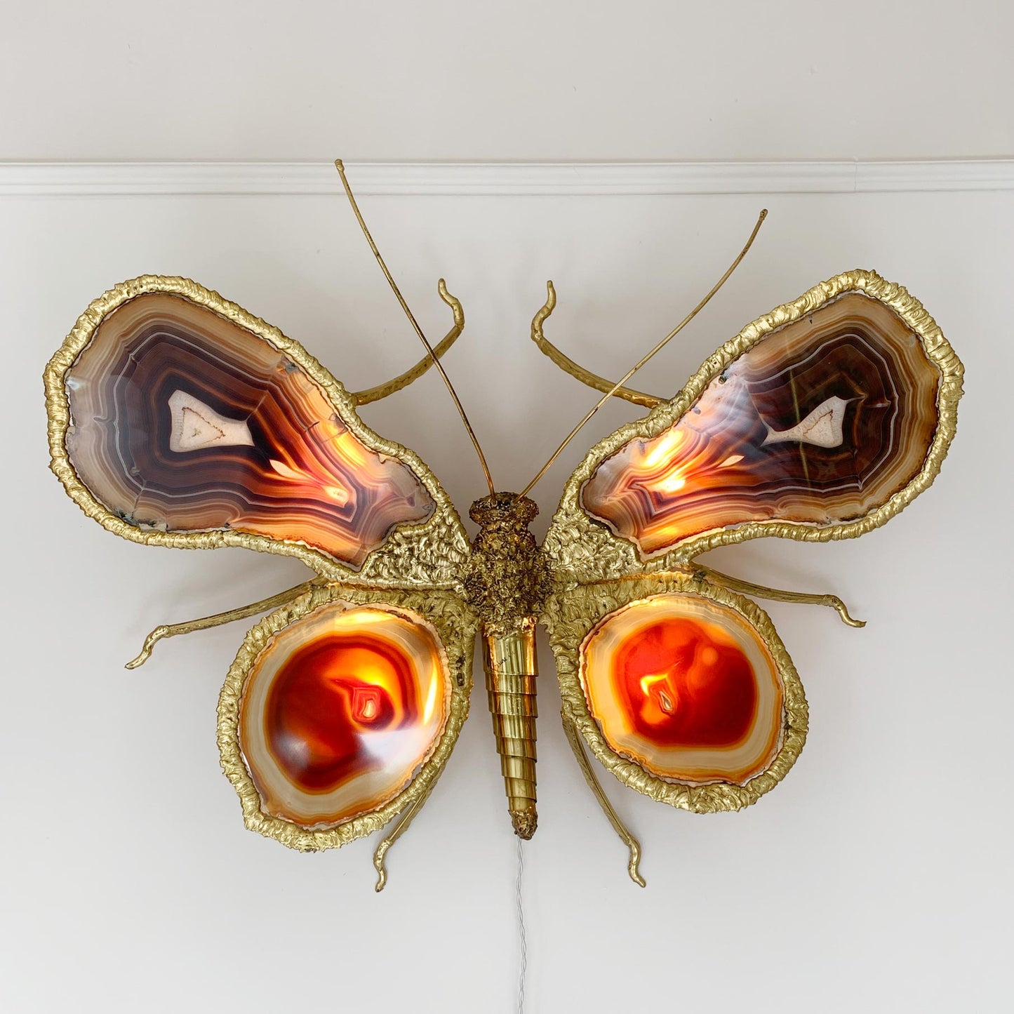 A decorative butterfly wall sculpture by Isabelle Faure,  made from gold-toned metal and polished agate slices, mounted on a white wall. The wings show layered natural agate patterns in brown, cream, and orange, with glowing warm light inside the lower wing sections.
