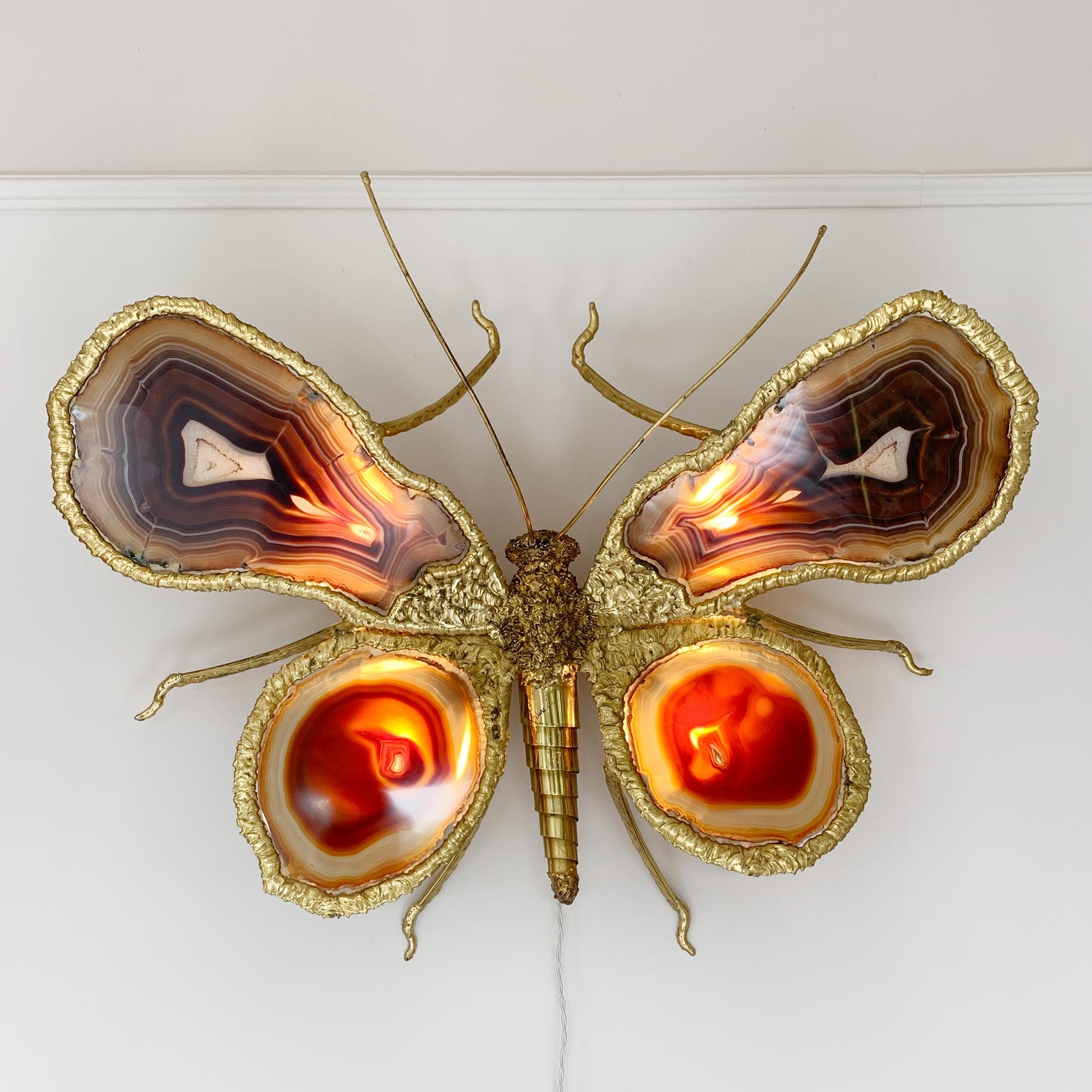 A decorative butterfly wall sculpture by Isabelle Faure,  made from gold-toned metal and polished agate slices, mounted on a white wall. The wings show layered natural agate patterns in brown, cream, and orange, with glowing warm light inside the lower wing sections.