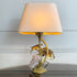 A decorative Isabelle Faure table lamp with a warm beige textured fabric shade, a gold-finished metal base sculpted as grape leaves and stems, and a cluster of pale pink quartz grapes, shown lit on a dark reflective surface against a light gray paneled wall.