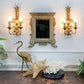 An ornate antique Italian gold-framed mirror is mounted on a white wall between two matching gold pineapple-style wall sconces. Below, a dark tabletop holds a gold crane figurine, a beige starfish, and a white coral arrangement on a black base.