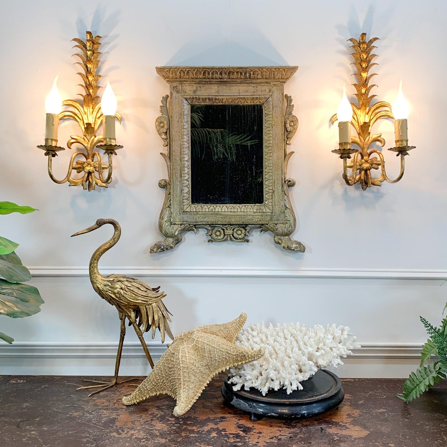 An ornate antique Italian gold-framed mirror is mounted on a white wall between two matching gold pineapple-style wall sconces. Below, a dark tabletop holds a gold crane figurine, a beige starfish, and a white coral arrangement on a black base.
