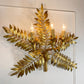 A gold-finished metal wall sconce designed as a fern leaf cluster with three lit candle-style bulbs, mounted on a white wall.