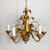 A vintage six-light chandelier with a gold leaf metal body, curved scrollwork, and dark brown wooden candle-style arms with white candle sleeves.