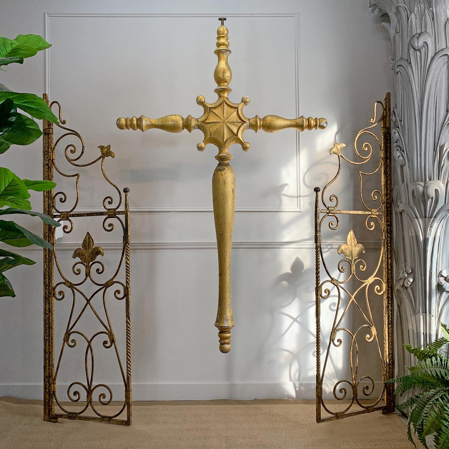 A decorative gold-toned wall cross hangs centered between two matching ornate wrought-iron gate panels with scrollwork and fleur-de-lis motifs, set against a white paneled wall with plants at the sides.