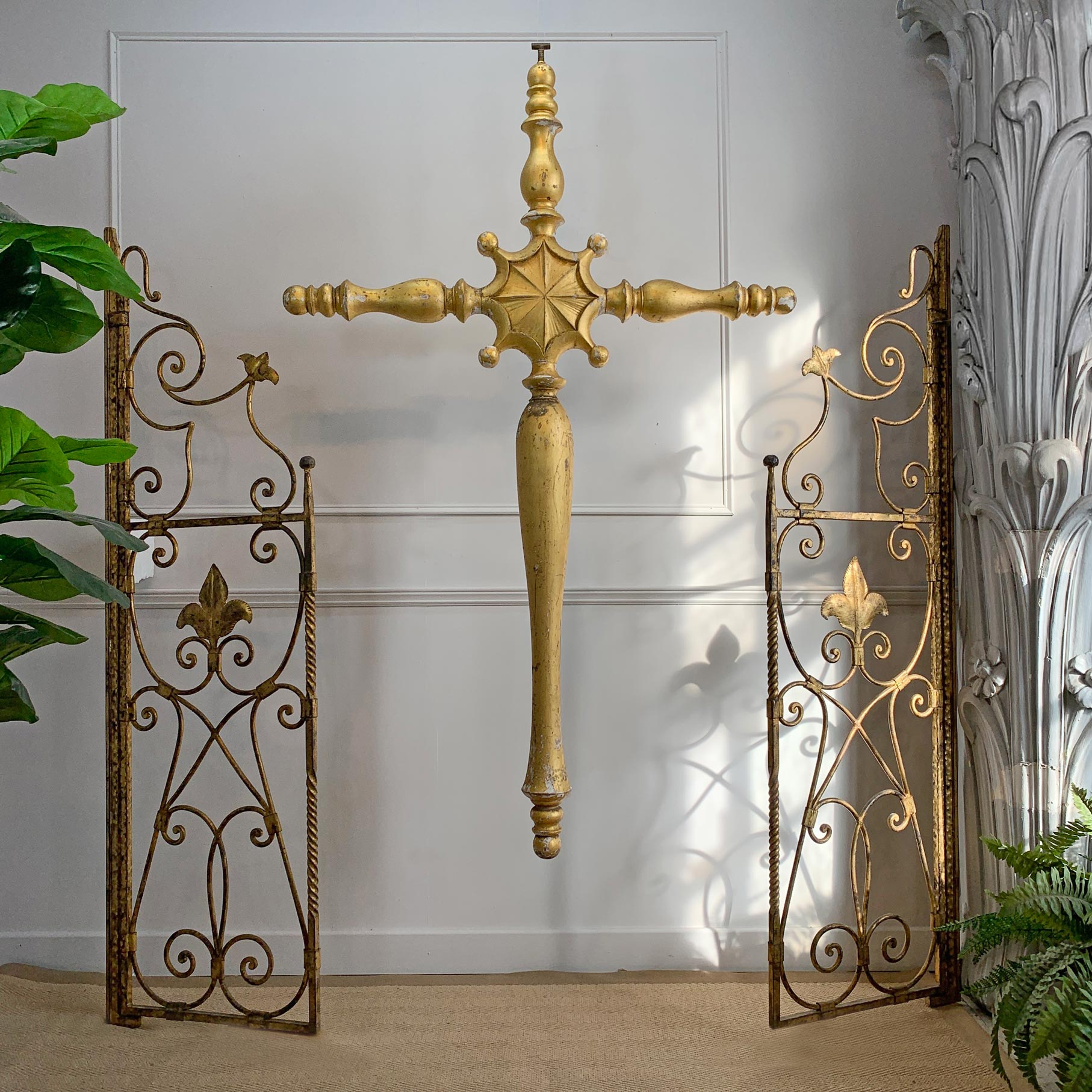 A decorative gold-toned wall cross hangs centered between two matching ornate wrought-iron gate panels with scrollwork and fleur-de-lis motifs, set against a white paneled wall with plants at the sides.