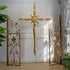 A decorative gold-toned wall cross hangs centered between two matching ornate wrought-iron gate panels with scrollwork and fleur-de-lis motifs, set against a white paneled wall with plants at the sides.