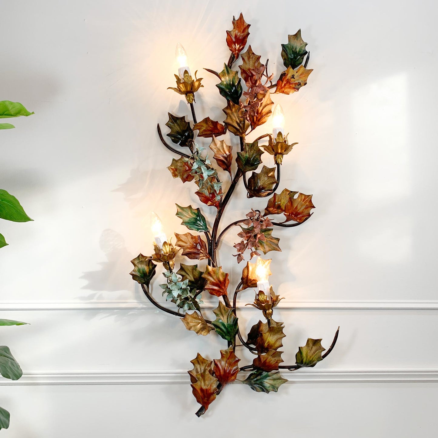 A decorative wall-mounted metal wall light with autumn holly leaf and vine flowers, in autumnal colours, lit with multiple warm candle-style bulbs against a white wall.