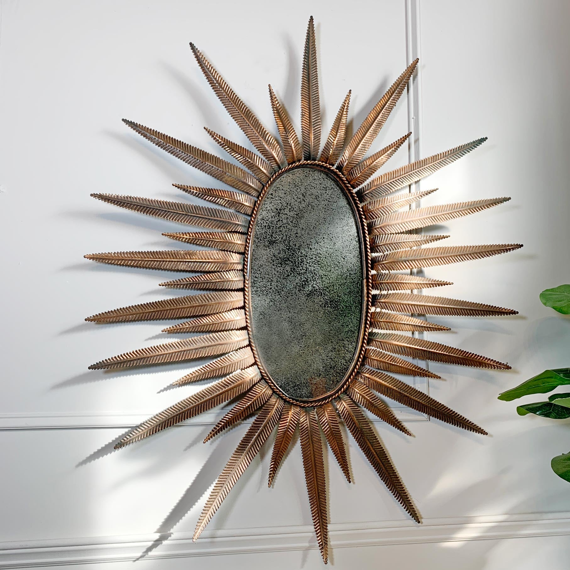 A decorative oval copper mirror with a sunburst frame in a textured metallic copper finish, mounted on a white paneled wall.