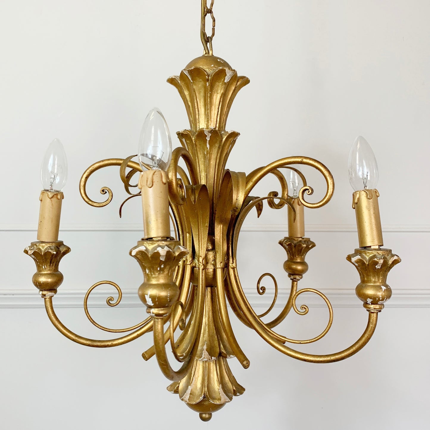 An ornate five-light brass chandelier with a classic baroque design, featuring sculpted floral elements and candle-style bulbs, shown against a plain white wall.