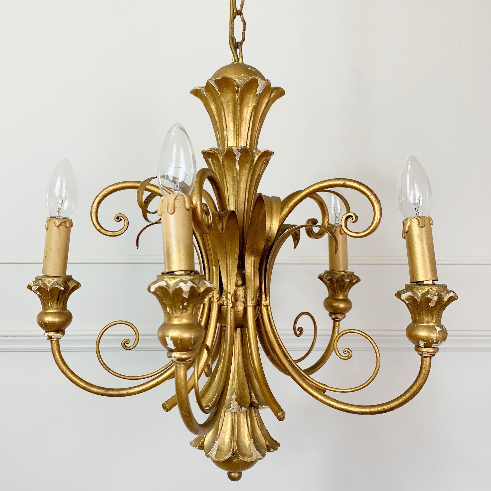 An ornate five-light brass chandelier with a classic baroque design, featuring sculpted floral elements and candle-style bulbs, shown against a plain white wall.