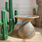 A sculptural woven side table with a twisted pedestal base and a circular glass top, wrapped in multicolored striped raffia. It is styled in a room setting next to green cactus decorations.