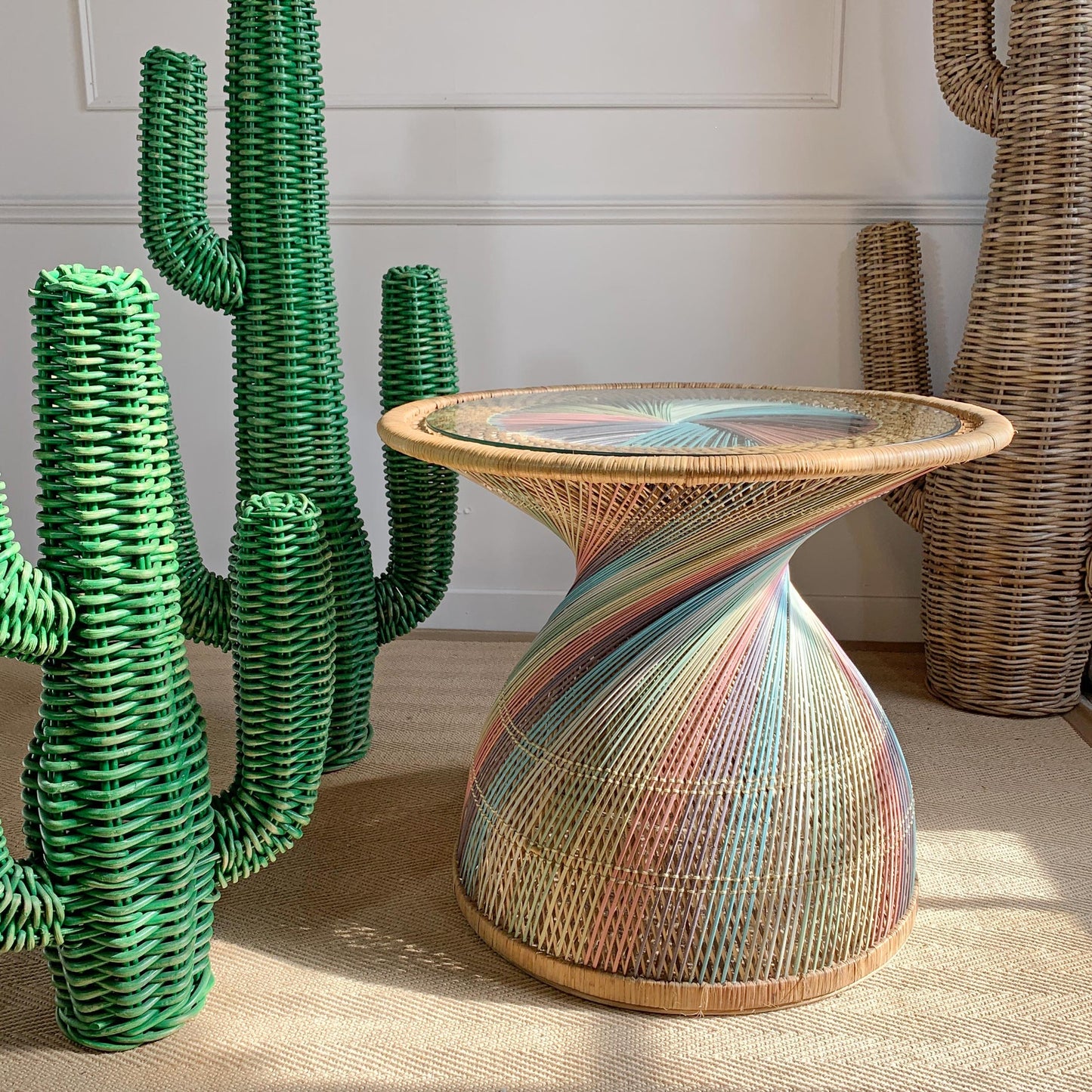 A sculptural woven side table with a twisted pedestal base and a circular glass top, wrapped in multicolored striped raffia. It is styled in a room setting next to green cactus decorations.