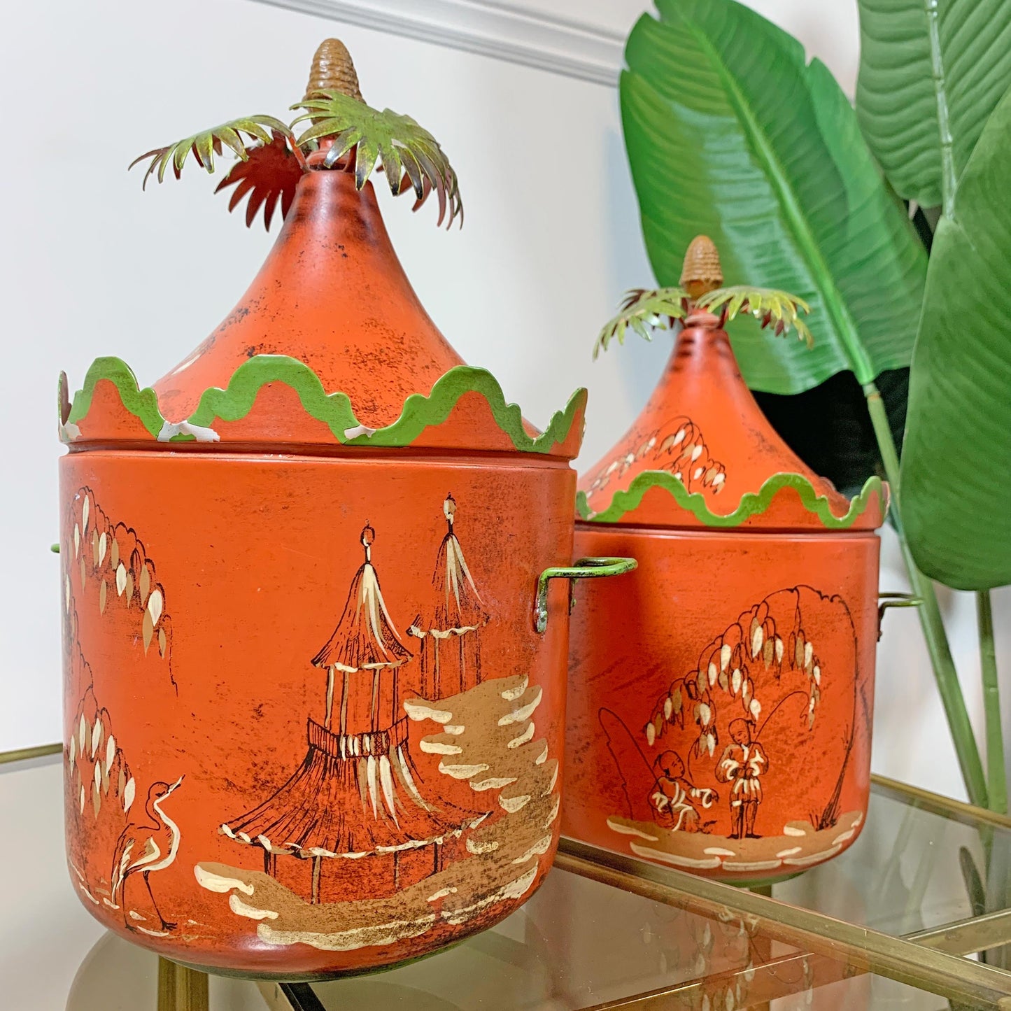 Two orange red decorative metal chinoiserie ice buckets with pointed lids and palm tree toppers, decorated with hand-painted beige and green chinoiserie patterns, displayed on a glass table with a tropical plant in the background.