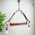 Industrial-style triangular pendant light fixture with metal frame, brown leather straps, and two hanging Edison bulbs, shown against a white wall with a plant at the side.