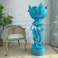 antique jerome massier turquoise blue ceramic lotus-shaped pedestal planter with sculpted figures on the base, displayed in an indoor room next to a distressed green chair and a potted plant.