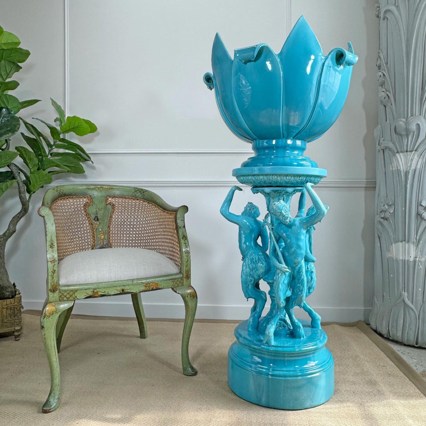 antique jerome massier turquoise blue ceramic lotus-shaped pedestal planter with sculpted figures on the base, displayed in an indoor room next to a distressed green chair and a potted plant.