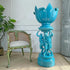 antique jerome massier turquoise blue ceramic lotus-shaped pedestal planter with sculpted figures on the base, displayed in an indoor room next to a distressed green chair and a potted plant.