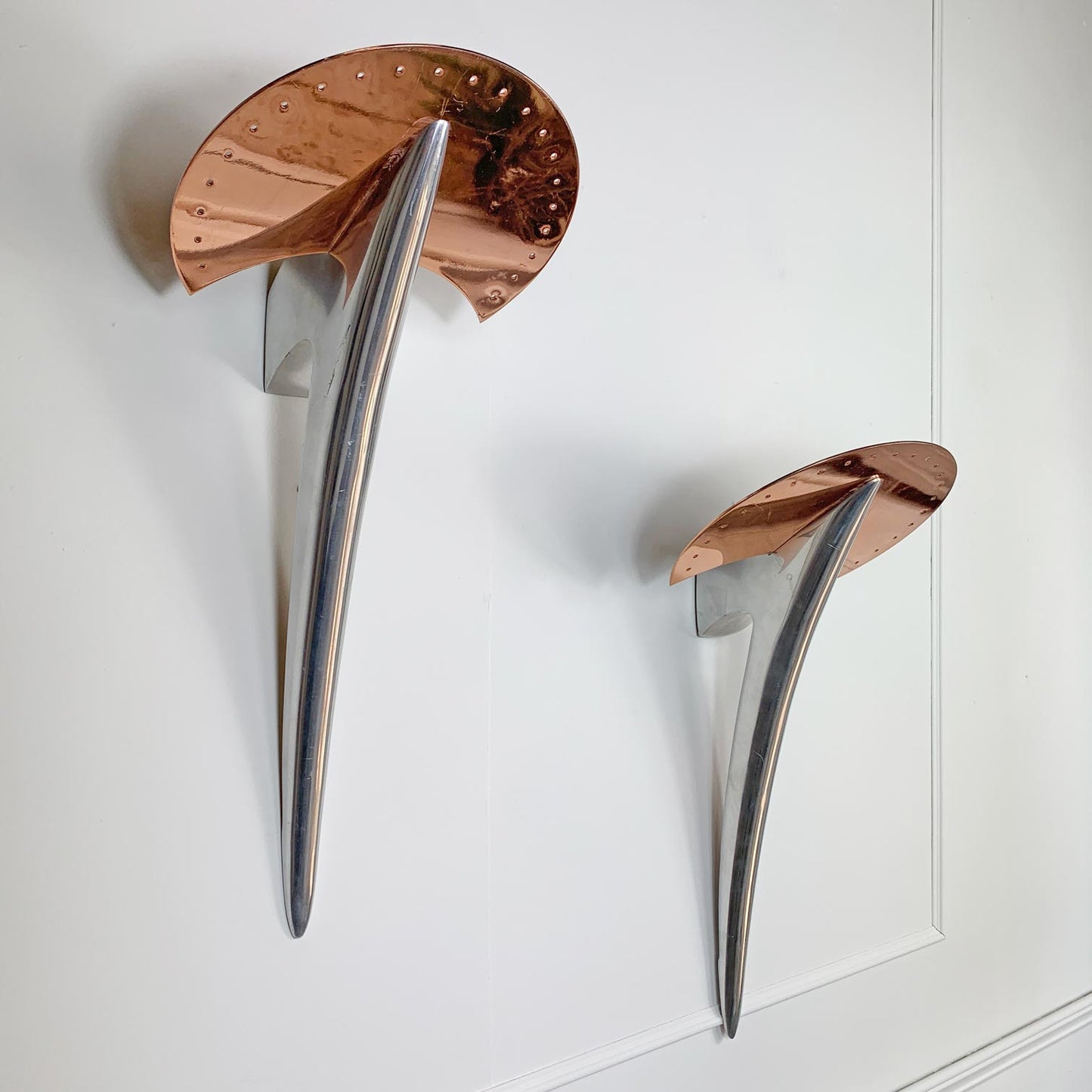 Two vintage wall sconces with reflective copper and polished silver metal leaf-like shapes, mounted on a white paneled wall.