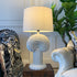 A sculptural white cactus shape table lamp with a textured, spiky tree-like base and a cream fabric drum shade, styled on a dark wooden side table between two upholstered armchairs in a living room setting.