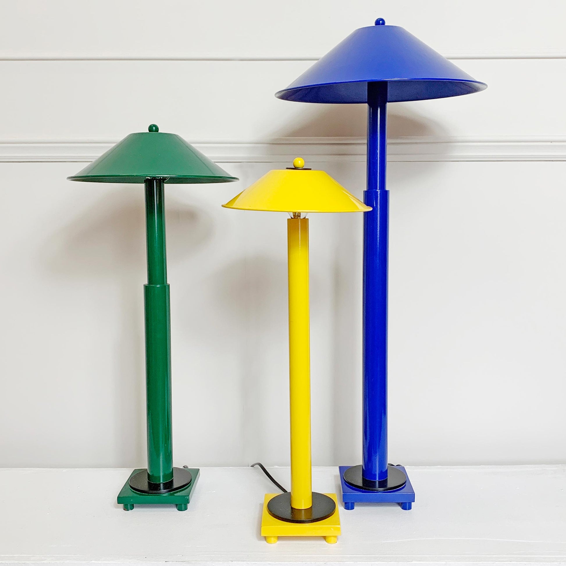 Three colorful metal table lamps with conical shades and cylindrical stems, shown in green, yellow, and blue on a white surface against a white paneled wall.