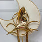 A close-up of a decorative gold-plated metal table lamp with sculptural leaf-like branches and a large cream-colored circular shade with a gold rim, mounted against a white paneled wall.
