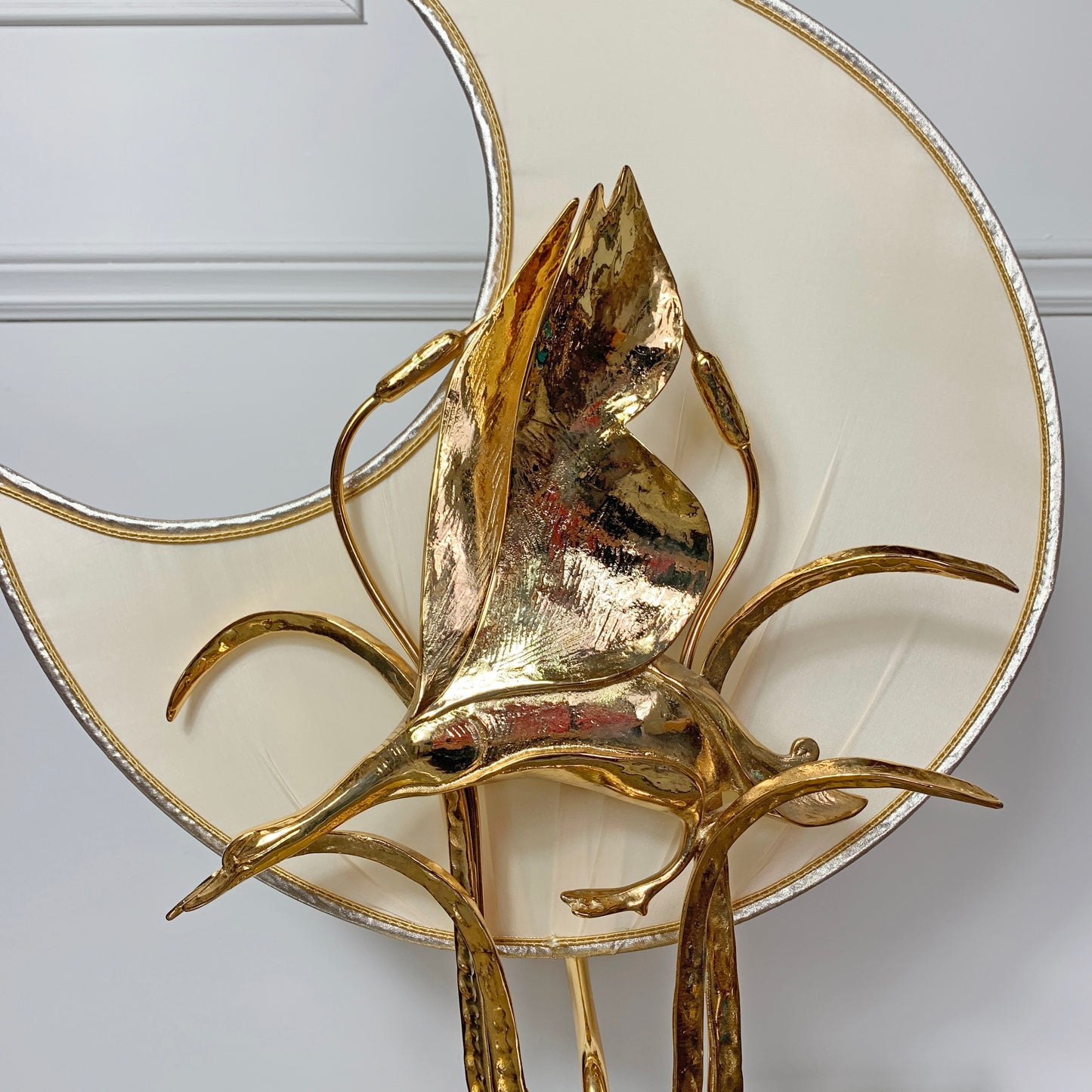 A close-up of a decorative gold-plated metal table lamp with sculptural leaf-like branches and a large cream-colored circular shade with a gold rim, mounted against a white paneled wall.