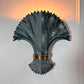 A decorative wall sconce with a sculptural fan or palm leaf design in dark charcoal gray, accented by a metallic gold band near the base, mounted on a white wall with warm light glowing from behind.