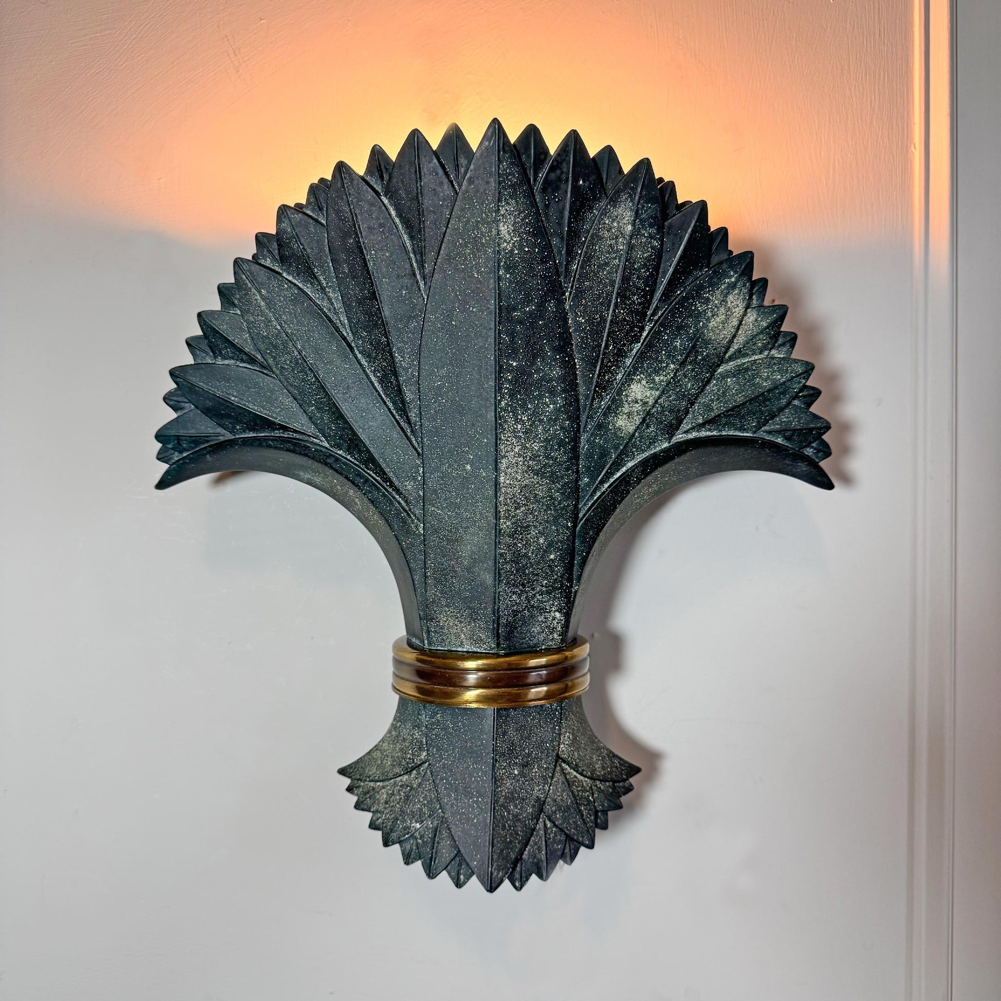 A decorative wall sconce with a sculptural fan or palm leaf design in dark charcoal gray, accented by a metallic gold band near the base, mounted on a white wall with warm light glowing from behind.