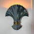 A decorative wall sconce with a sculptural fan or palm leaf design in dark charcoal gray, accented by a metallic gold band near the base, mounted on a white wall with warm light glowing from behind.