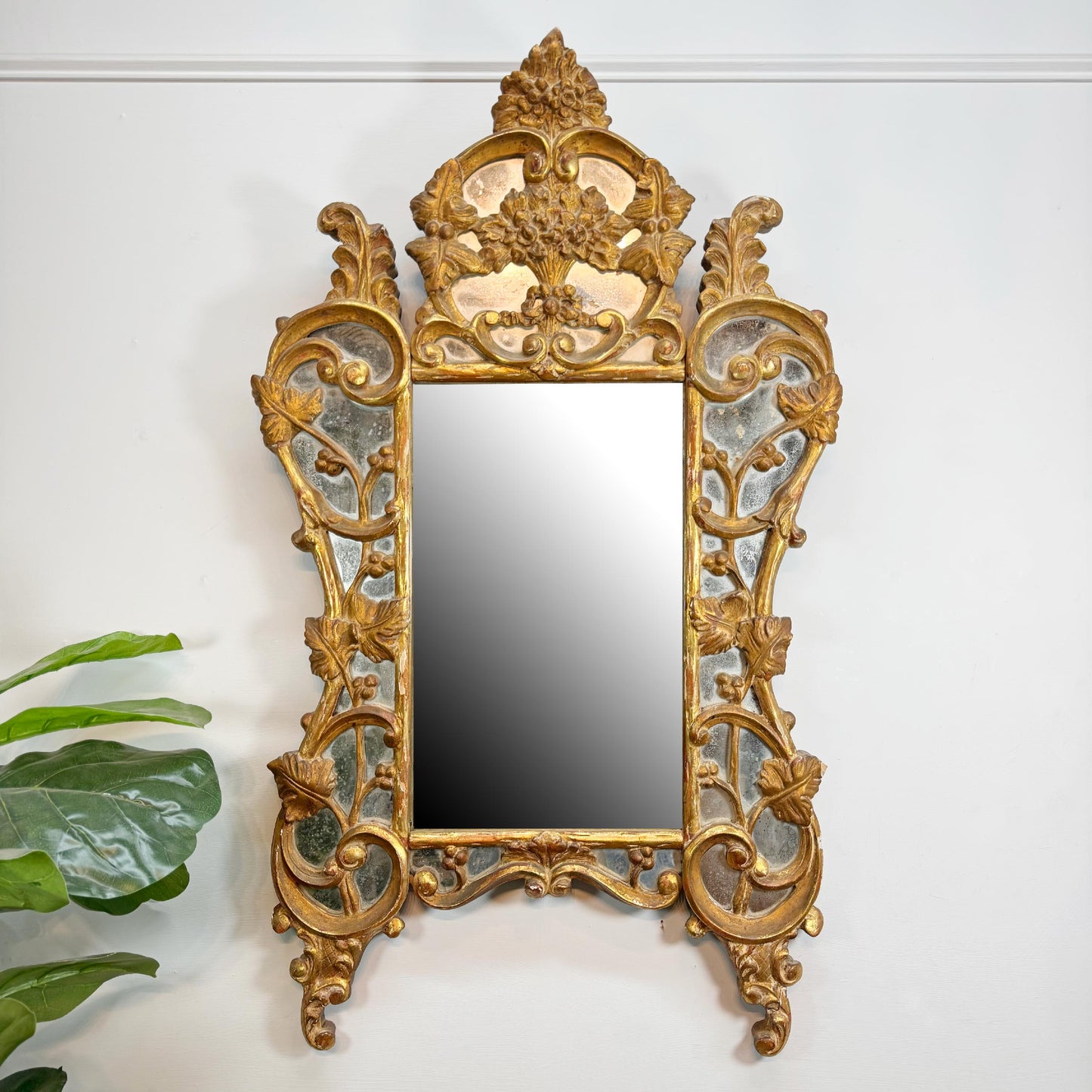 An ornate antique wall mirror with a gilded gold frame featuring intricate floral carvings and a central rectangular mirror, mounted on a white wall with a green plant leaf visible in the lower left corner.