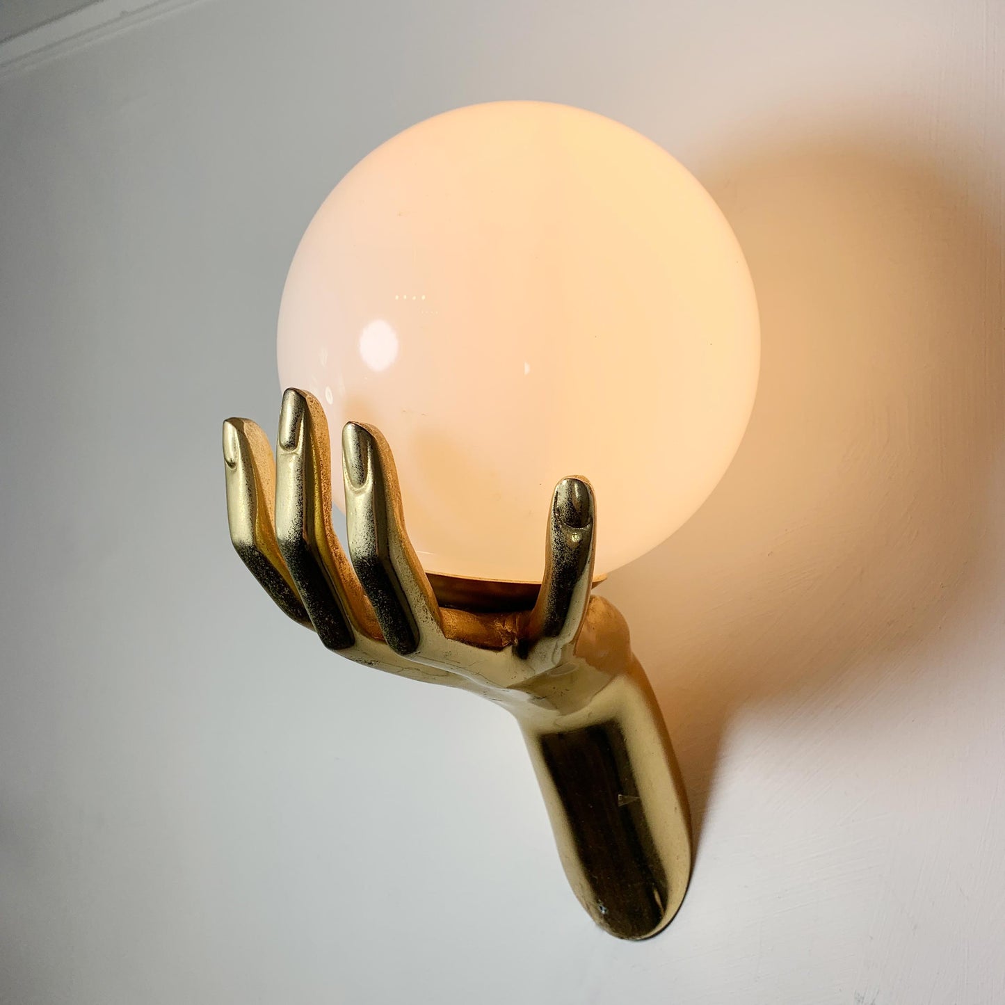 A wall-mounted decorative bronze light fixture shaped like a metallic gold hand holding a large glowing frosted globe shade, shown against a plain light wall.