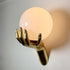 A wall-mounted decorative bronze light fixture shaped like a metallic gold hand holding a large glowing frosted globe shade, shown against a plain light wall.