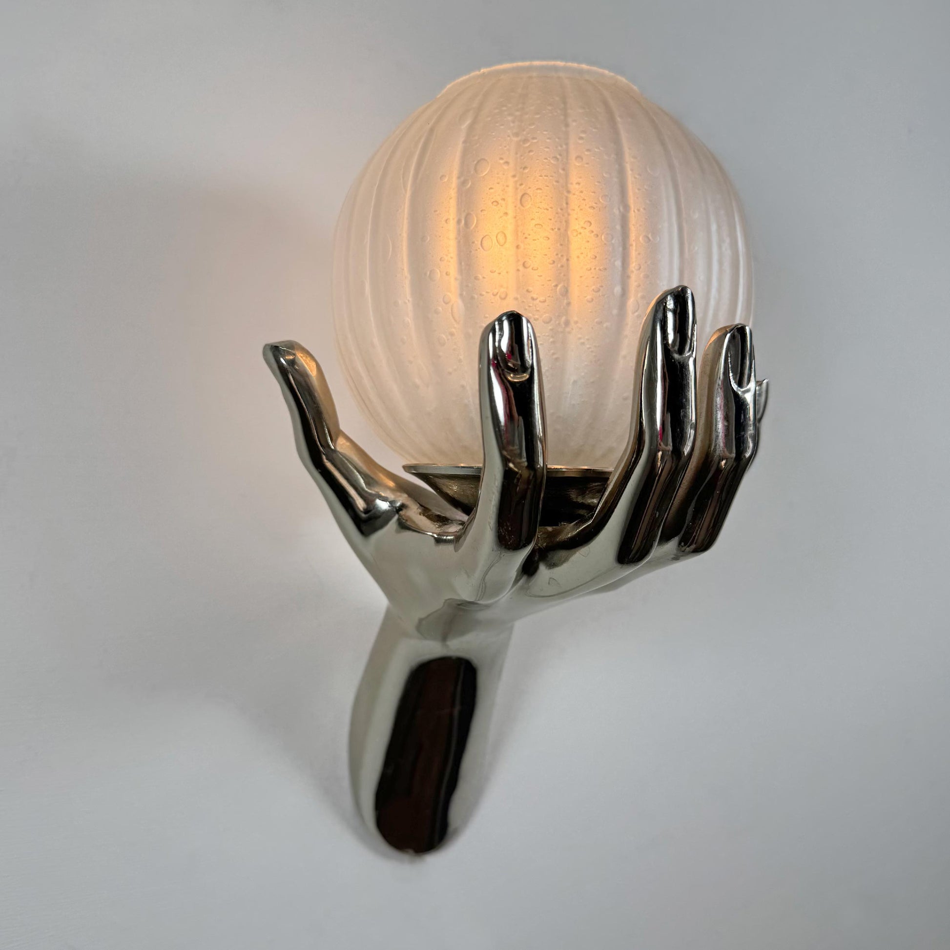 A sculptural Maiosn Arlus Hand wall sconce featuring a polished chrome hand holding a ribbed, frosted glass globe shade that glows with a warm light, set against a plain light gray background.