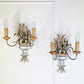A Pair of Maison Bagues Gilt Crystal Wall Lights with curved metal larms, candle-shaped bulbs, feather-like decorative elements, and a clear glass bowl base, shown against a white paneled wall.