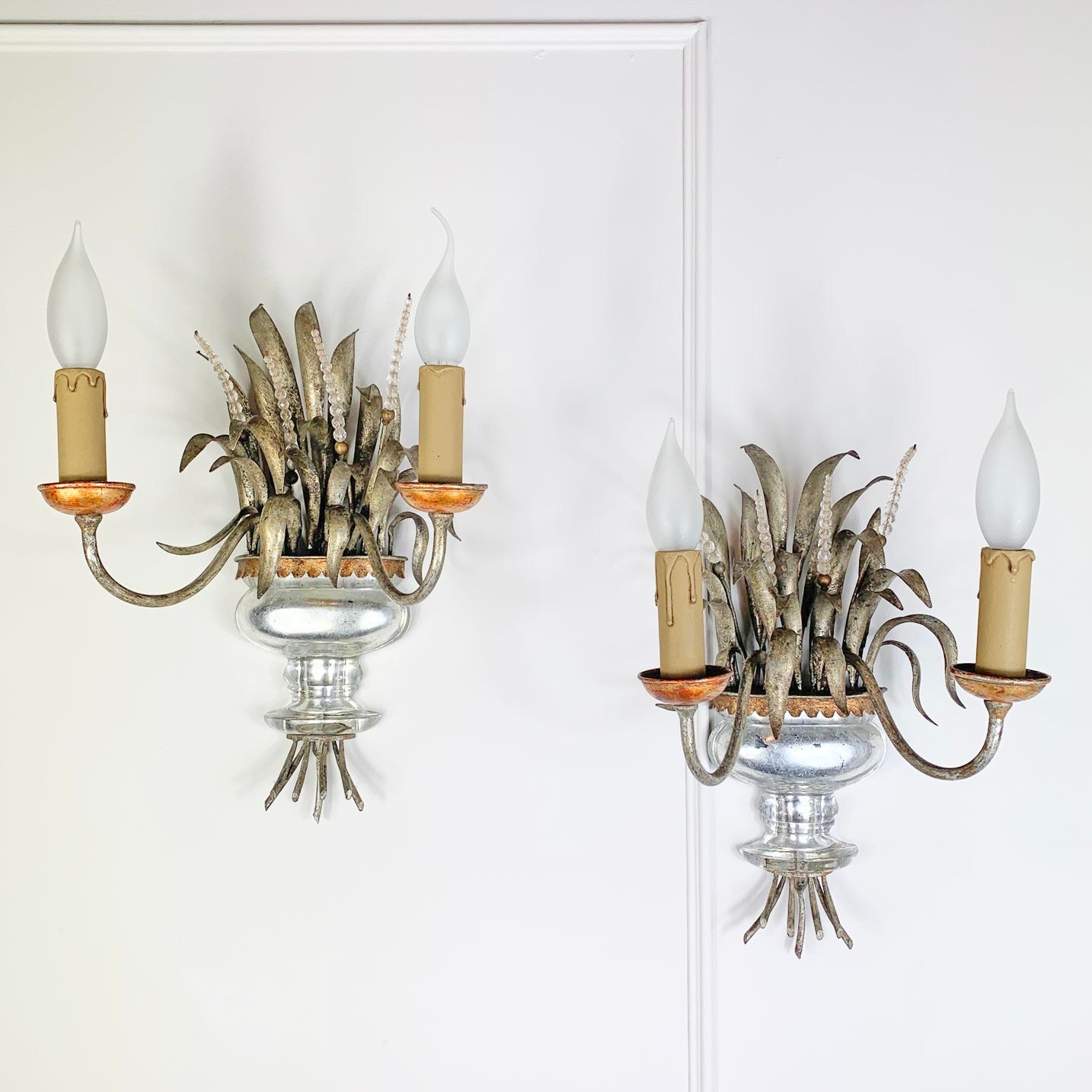 A Pair of Maison Bagues Gilt Crystal Wall Lights with curved metal larms, candle-shaped bulbs, feather-like decorative elements, and a clear glass bowl base, shown against a white paneled wall.