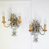 A Pair of Maison Bagues Gilt Crystal Wall Lights with curved metal larms, candle-shaped bulbs, feather-like decorative elements, and a clear glass bowl base, shown against a white paneled wall.